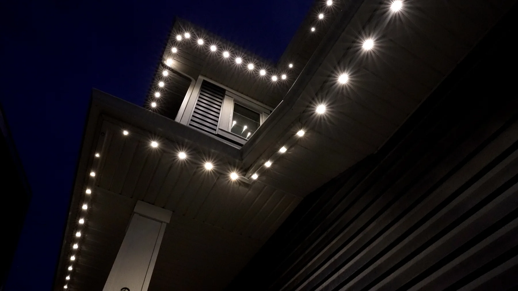 Looking up at a soffit at night, white emitters evenly spaced along track fitted tight under the eave and around a dormer.
